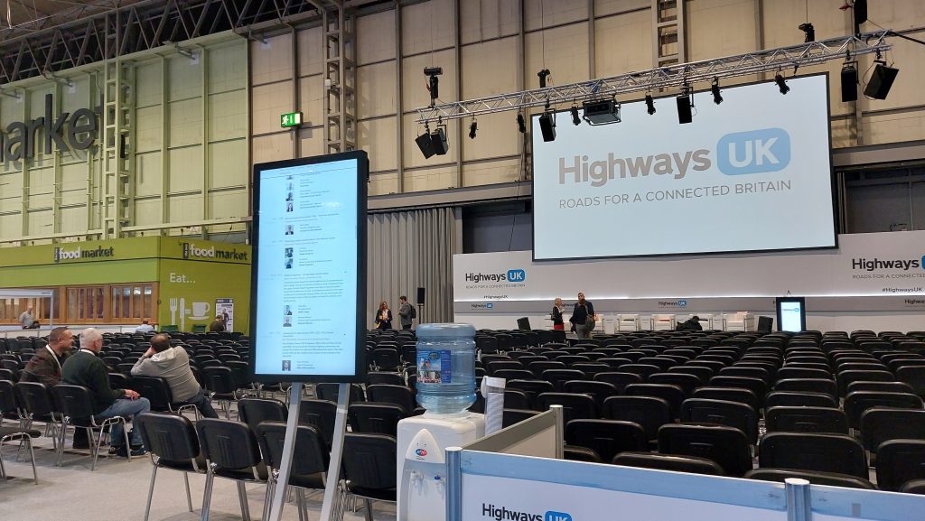 The main conference stage for Highways UK at the Highways UK event. On the right is a large screen displaying the Highways UK logo UK. Facing the screen is a row of chairs for attendees to sit 