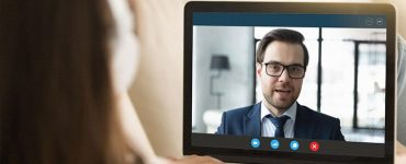 A woman wearing headphones is looking at a computer screen where she is having a video interview with a man who is wearing a suit.