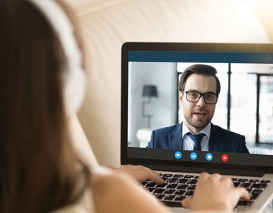 A woman wearing headphones is looking at a computer screen where she is having a video interview with a man who is wearing a suit.