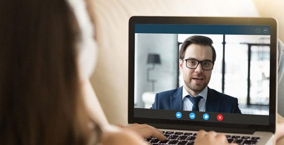 A woman wearing headphones is looking at a computer screen where she is having a video interview with a man who is wearing a suit.